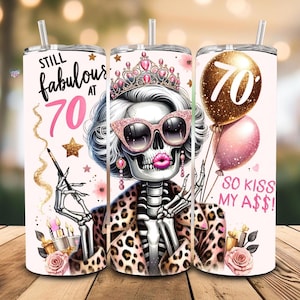 Op de afbeelding: Een witte tumbler met een roze en gouden ontwerp. Het ontwerp toont een skelet dat een luipaardprintjasje en een kroon draagt. De tekst "Still Fabulous at 70" staat aan de linkerkant van de tumbler. De tekst "So Kiss My A$$!" staat aan de rechterkant van de tumbler.