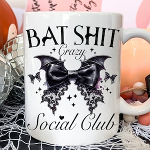 May include: White ceramic mug with a black graphic that says "Bat Shit Crazy Social Club". The graphic features a black bow with bat wings and lace trim.
