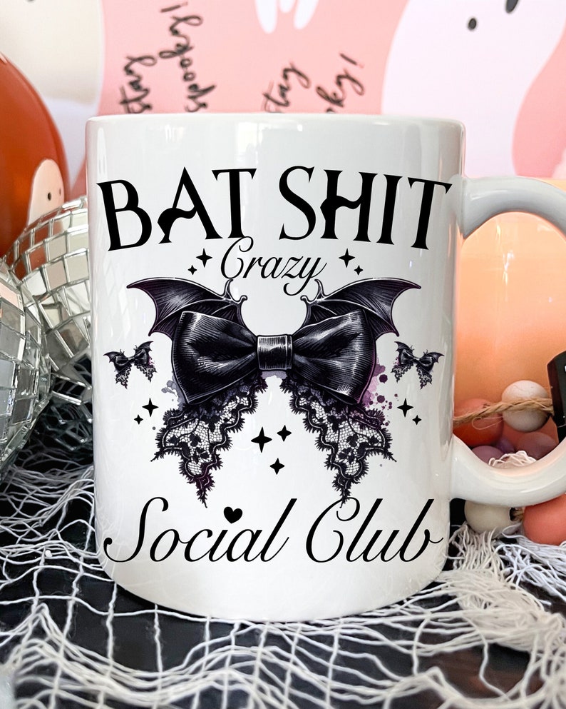 May include: White ceramic mug with a black graphic that says "Bat Shit Crazy Social Club". The graphic features a black bow with bat wings and lace trim.