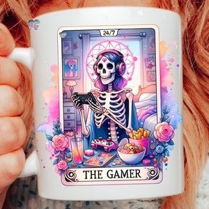 K&ouml;nnte beinhalten: Eine wei&szlig;e Keramiktasse mit einer bunten Illustration eines Skeletts, das Kopfh&ouml;rer tr&auml;gt und einen Videospielcontroller h&auml;lt. Der Text "24/7 THE GAMER" ist auf die Tasse gedruckt.