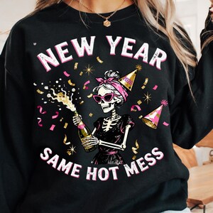 Op de afbeelding: Zwarte sweatshirt met de tekst "NEW YEAR SAME HOT MESS". De afbeelding toont een skelet met een zonnebril, een roze haarband en een feesthoedje, die een champagnefles vasthoudt. Confetti en roze accenten maken het ontwerp compleet.