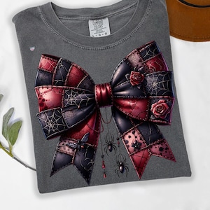 以下が含まれることがあります： 黒と赤の大きなリボン模様が特徴のグレーのComfort Colors Tシャツ。クモの巣、バラ、ぶら下がっているクモで飾られています。シャツはクルーネックで、「Comfort Colors」と書かれたタグが付いています。
