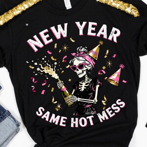 Op de afbeelding: Zwart T-shirt met een skeletafbeelding met een zonnebril en een feesthoedje, die een champagnefles ontkurkt. De tekst luidt "NEW YEAR SAME HOT MESS" in roze en wit.