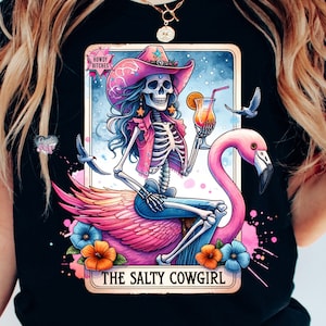 May include: A black t-shirt with a colorful illustration of a skeleton cowgirl riding a pink flamingo. The illustration is in the style of a tarot card and includes the text "Howdy Bitches" and "The Salty Cowgirl".