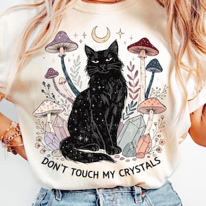 Könnte beinhalten: Cremefarbenes T-Shirt mit einem schwarzen Kater-Design, Sternen, Pilzen, Kristallen und dem Text "DON'T TOUCH MY CRYSTALS". Das Design beinhaltet einen Mond und Sterne.