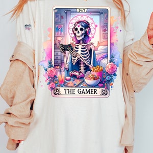 K&ouml;nnte beinhalten: Ein wei&szlig;es T-Shirt mit einer bunten Illustration eines Skeletts, das Videospiele spielt. Die Illustration ist im Stil einer Tarotkarte und enth&auml;lt den Text "The Gamer" und "24/7".