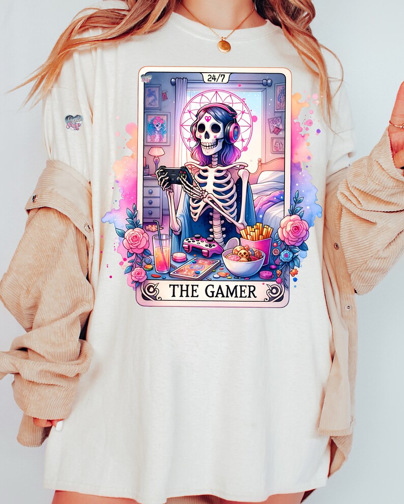 K&ouml;nnte beinhalten: Ein wei&szlig;es T-Shirt mit einer bunten Illustration eines Skeletts, das Videospiele spielt. Die Illustration ist im Stil einer Tarotkarte und enth&auml;lt den Text "The Gamer" und "24/7".