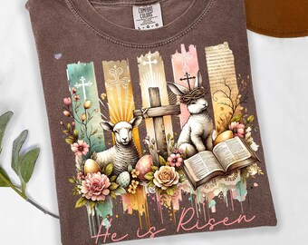 May include: A brown t-shirt with a watercolour design featuring a lamb, a rabbit, and a cross. The design includes flowers, eggs, and the words "He is Risen".