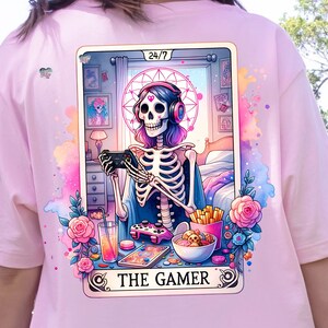 K&ouml;nnte beinhalten: Eine Tarotkarten-Illustration mit einem Skelett, das Kopfh&ouml;rer tr&auml;gt und einen Videospiel-Controller h&auml;lt. Die Karte tr&auml;gt den Titel "The Gamer" und hat ein 24/7-Symbol in der oberen rechten Ecke. Der Hintergrund ist ein rosa-blaues Aquarelldesign mit Blumen und anderen spielbezogenen Bildern.