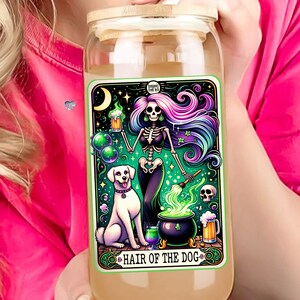 May include: A glass jar with a wooden lid and a colorful illustration of a skeleton holding a beer mug and a cauldron with green smoke. The illustration is surrounded by a green border and the text "Hair of the Dog" is at the bottom.