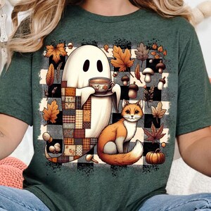 May include: Green t-shirt with an autumnal design. A ghost holds a teacup, alongside a fox, autumn leaves, mushrooms, and a pumpkin. The background features a black and white checkered pattern.