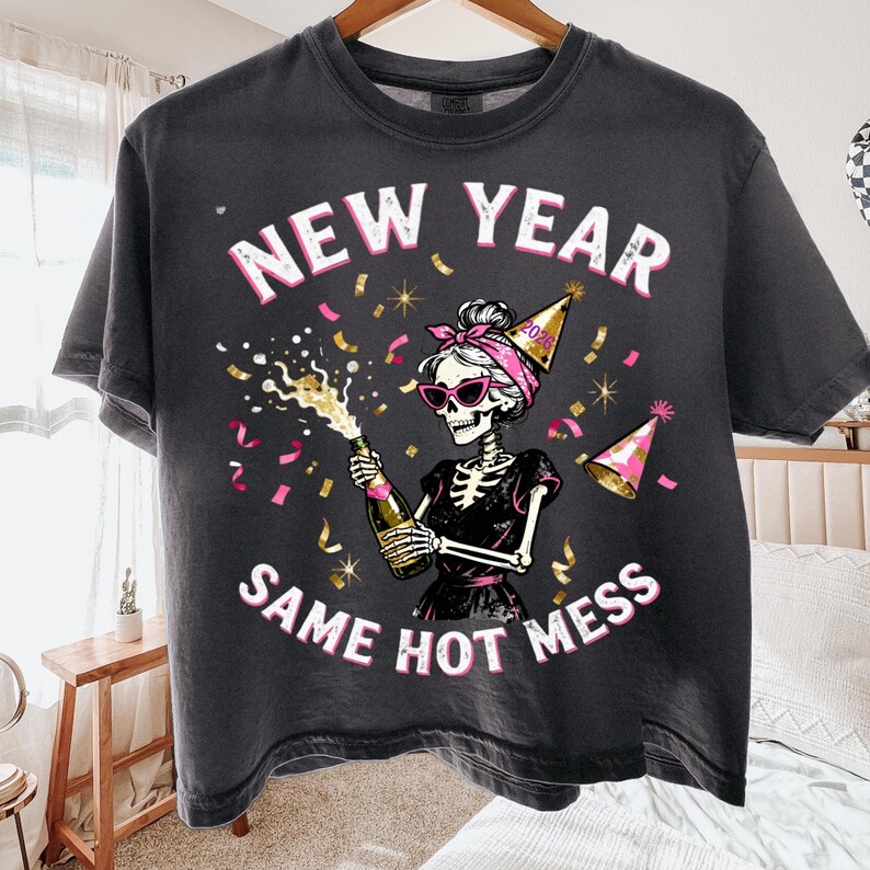 Op de afbeelding: Donkergrijs T-shirt met de tekst "NEW YEAR SAME HOT MESS" in roze en wit. De afbeelding toont een skelet met een zonnebril, een roze haarband en een feesthoed, die een champagnefles vasthoudt. Confetti is verspreid rond het ontwerp.