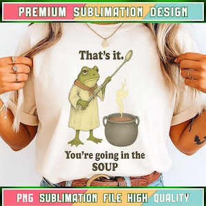 Op de afbeelding: Crèmekleurig T-shirt met een cartoon kikkerkok die een lepel boven een soeppot vasthoudt. De tekst "That's it. You're going in the SOUP" staat onder de kikker. Het shirt heeft groene en roze tekst aan de boven- en onderkant.