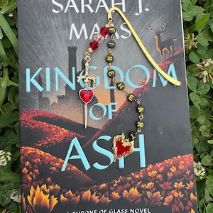 Op de afbeelding: Een boek getiteld "Kingdom of Ash" van Sarah J. Maas met een gouden bladwijzer en een kralenbedel. De bedel heeft een zwaard, een hart en het woord "Fireheart". Het boek ligt op een bed van groen.