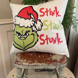 May include: A square off-white throw pillow featuring a graphic of a green character wearing a red and white Santa hat. The text Stink. Stank. Stunk. is printed in red and light green lettering on the right side of the pillow cover fabric.