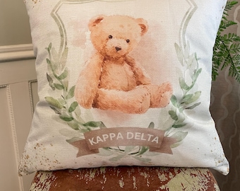 May include: A square throw pillow featuring a light beige fabric with a centered illustration of a tan teddy bear. The bear is framed by green leafy branches and a brown ribbon banner with the white text KAPPA DELTA printed on it.