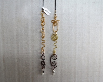 May include: A pair of handcrafted wire earrings. One earring is gold-toned with a wavy design and a dark brown spiral at the bottom. The other earring is gold and yellow-toned with a spiral and wavy design. Both have small clear beads at the bottom.