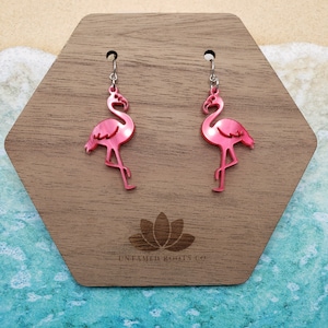 May include: A pair of pink flamingo earrings with silver-colored hooks. The earrings are displayed on a wooden, hexagonal stand. The stand has a lotus flower design and the text "UNFAMED ROOTS CO" etched on it.