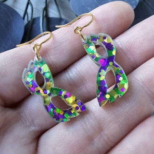 May include: A pair of gold dangle earrings with a clear resin mask shape filled with iridescent green, purple, and gold glitter.