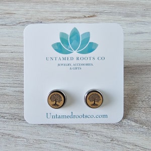 Pode incluir: Um par de brincos de press&atilde;o de prata com uma incrusta&ccedil;&atilde;o de madeira que apresenta um design de &aacute;rvore da vida. Os brincos s&atilde;o exibidos em um cart&atilde;o branco com o texto "Untamed Roots Co. Joias, Acess&oacute;rios e Presentes" e "Untamedrootsco.com".