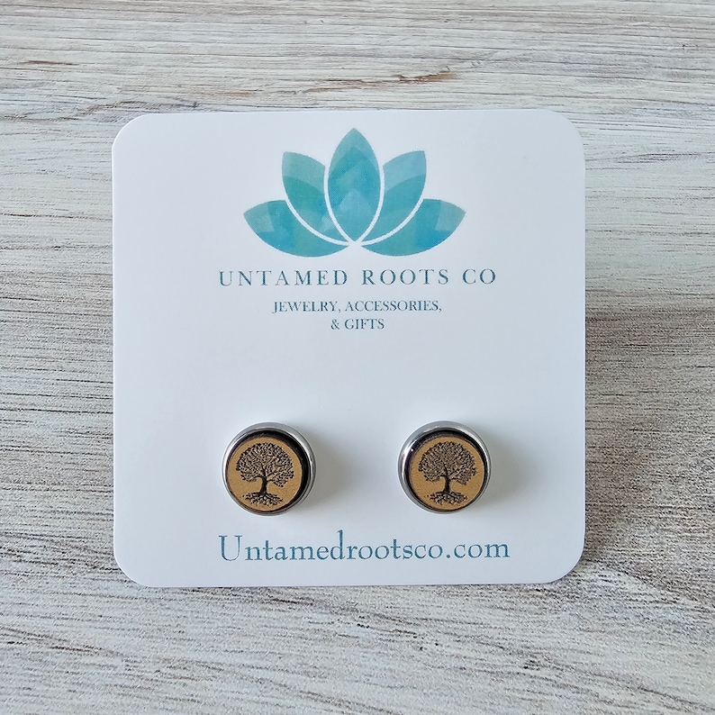 Pode incluir: Um par de brincos de press&atilde;o de prata com uma incrusta&ccedil;&atilde;o de madeira que apresenta um design de &aacute;rvore da vida. Os brincos s&atilde;o exibidos em um cart&atilde;o branco com o texto "Untamed Roots Co. Joias, Acess&oacute;rios e Presentes" e "Untamedrootsco.com".