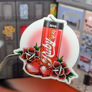 May include: A red and white can of Ruby Zero Sugar soda with a red devil illustration and the text "Ruby" in a stylized font. The can is surrounded by two red roses and two red hearts.
