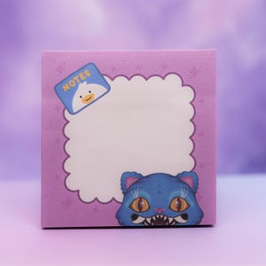 May include: A square notepad with a purple background and a white, cloud-shaped space for notes. The notepad features a blue cat illustration at the bottom and a small square with the word "NOTES" and a duck illustration.