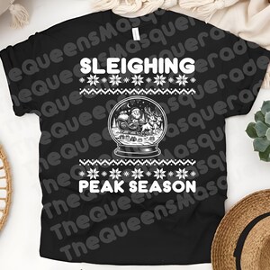 May include: Black t-shirt with a festive design. The shirt features the words "SLEIGHING" and "PEAK SEASON" in white, along with a snow globe graphic of Santa Claus and reindeer. A winter holiday theme.