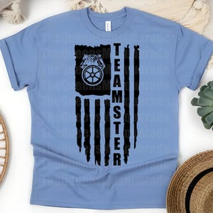 May include: Light blue t-shirt featuring a distressed black graphic resembling an American flag. The graphic includes a horse-drawn carriage wheel and the word "TEAMSTER" in bold, vertical letters. The shirt is made of soft material.