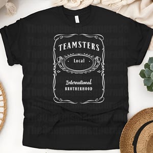 May include: Black t-shirt with a white graphic design. The design features the text "TEAMSTERS Local International Brotherhood" in a vintage-style frame. The shirt is displayed on a white surface with a straw hat and a succulent plant.
