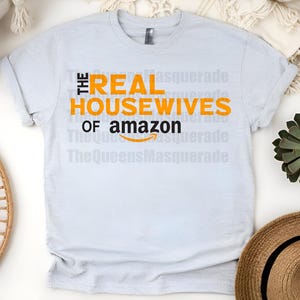 May include: A light blue t-shirt with the text "THE REAL HOUSEWIVES OF amazon" in orange and black. The Amazon logo is featured below the text. The shirt is displayed on a white surface with decorative items.