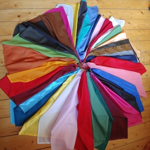Könnte beinhalten: Eine Sammlung farbenfroher Seidenschals, kreisförmig angeordnet. Die Schals zeigen eine große Auswahl an Farben, darunter Rot, Blau, Gelb, Rosa, Grün und Braun. Der Stoff hat eine glänzende, glatte Textur, und die Schals liegen auf einer Holzoberfläche.