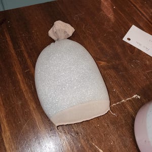 May include: A white, glitter-covered, egg-shaped object encased in a sheer, beige, stocking-like fabric. The fabric is gathered and tied at the top, creating a small, knotted protrusion, while the bottom edge features a stitched, beige hem.