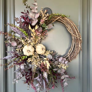 Peut inclure: Une couronne de saison sur une base en vigne, ornée de fausses citrouilles et d'une variété de feuillages artificiels aux tons bordeaux, beige et vert. Parfait pour la décoration automnale.