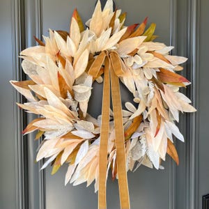Peut inclure: Une couronne décorative avec un design circulaire de feuilles superposées de couleur crème avec des touches d'orange et de vert. Un ruban doré est noué en haut, avec de longues queues flottantes. La couronne est affichée sur une porte grise.