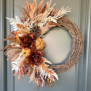 Peut inclure: Une couronne circulaire faite de brindilles brunes, ornée sur un côté d'herbes séchées, de feuillage blanc semblable à des fougères, de fleurs en tissu orange foncé, de deux petites citrouilles texturées et d'une gousse de lotus séchée.