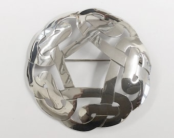 Pode incluir: Um broche de nó celta de prata com um design circular. O broche é feito de metal polido e tem um padrão intrincado de nós entrelaçados.