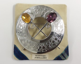 May include: Silver brooch with a Celtic knot design and three gemstones. The brooch has a yellow, purple, and clear gemstone. The brooch is on a white background with the text "Scottish Jewellery".
