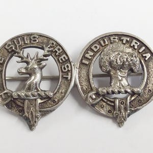 Puede incluir: Un broche plateado con dos diseños circulares. El lado izquierdo presenta un ciervo con el texto "Je Suis Prest" a su alrededor. El lado derecho presenta un árbol con el texto "Industria" a su alrededor.