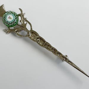 May include: A silver-toned metal kilt pin featuring a thistle design. The top of the pin is adorned with a round, green and white millefiori glass cabochon that displays a small purple thistle flower graphic in the center of the pattern.