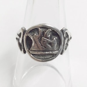 Peut inclure: Bague argentée avec un emblème circulaire détaillé. L'emblème présente un motif stylisé, peut-être un blason ou un symbole, avec des éléments en relief. La bague est posée sur un fond clair, mettant en valeur ses détails.