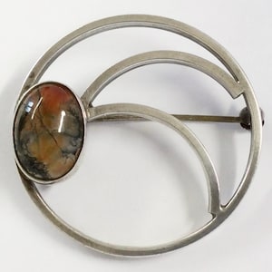 Peut inclure: Broche argentée avec une pierre ovale multicolore. La broche présente un design géométrique avec des lignes courbes et une fermeture à épingle. La pierre affiche des nuances de rouge, de brun et de noir.