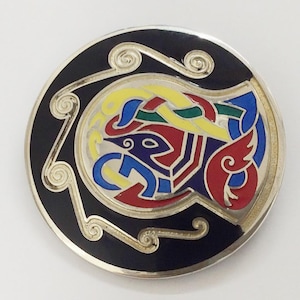 May include: A silver circular brooch with a black background and a colorful Celtic knot design. The knot design features a bird with red, blue, yellow, and green accents.