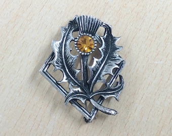May include: A silver-toned brooch featuring a thistle design with an amber-colored center stone. The brooch has a detailed, textured appearance, with a diamond-shaped frame. The thistle's leaves and petals are intricately carved.