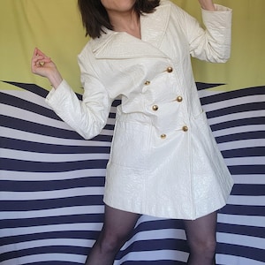 May include: A white, textured, double-breasted jacket with gold buttons. The jacket has a collar and a pocket on the left side. The jacket is worn over a black top and black tights.