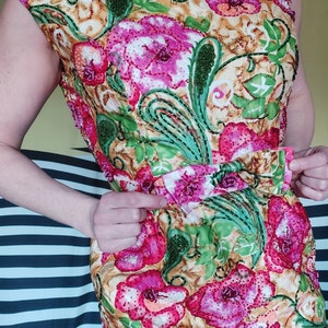 Op de afbeelding: Een mouwloze jurk met een bloemenprint in roze, groene en gele tinten. De jurk heeft een aansluitend bovenlijf en een zwierige rok. De stof is versierd met pailletten en kralen.