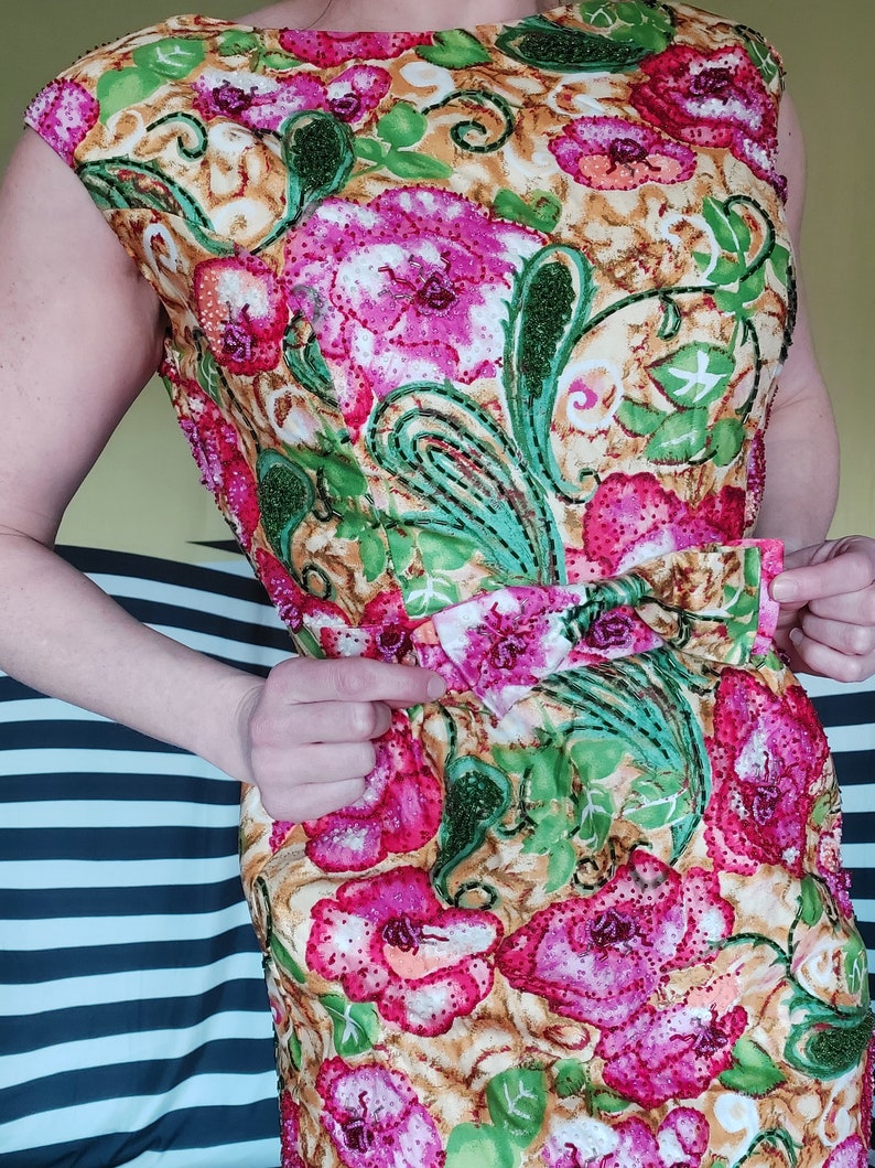 Op de afbeelding: Een mouwloze jurk met een bloemenprint in roze, groene en gele tinten. De jurk heeft een aansluitend bovenlijf en een zwierige rok. De stof is versierd met pailletten en kralen.