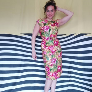 Op de afbeelding: Een vrouw draagt een jurk met bloemenprint met roze, groene en gele bloemen. De jurk heeft een aansluitende pasvorm en een ronde hals. Ze staat voor een gestreepte achtergrond.