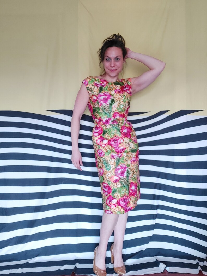 Op de afbeelding: Een vrouw draagt een jurk met bloemenprint met roze, groene en gele bloemen. De jurk heeft een aansluitende pasvorm en een ronde hals. Ze staat voor een gestreepte achtergrond.