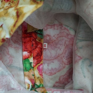 Op de afbeelding: Close-up van de binnenkant van een jurk met bloemenprint en een rode, groene en gouden bies. Het maatlabel geeft "14" aan.
